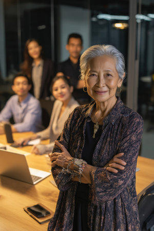 An elderly Asian businesswoman leans casually on a meeting table, arms folded with a confident smile, actively engaging her younger colleagues in creative discussion.の素材