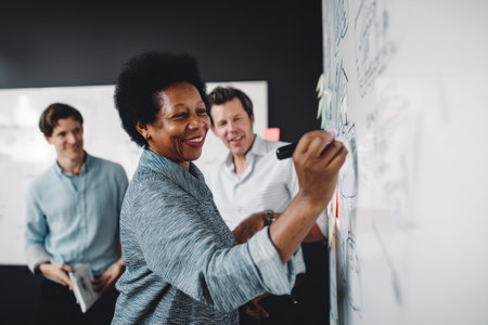 A middle-aged African-American woman collaborates on a whiteboard with marketing staff, refining branding messages and visual storytelling approaches for a new product campaign launchの素材