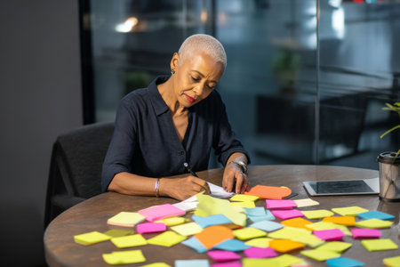 Elderly African-American businesswoman working on project planning at a round table, colorful sticky notes arranged neatly, emphasizing collaboration and senior experience in office cultureの素材