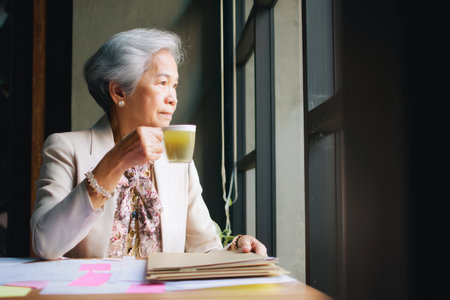 Elderly Asian businesswoman enjoying a quiet moment after a presentation, sipping green tea beside a window as paper slides rest in a neat folder. She's calm, accomplished, and stylish.の素材