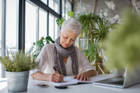 Elderly Caucasian business woman organizing her week with a digital calendar and hand-written notes, seated at her minimalist desk, wearing an elegant scarf and blouse combo, indoor plants and a large window fill the space with warmth and motivation for planningの素材