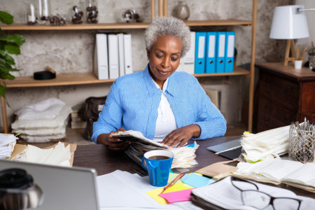 Mature African-American businesswoman preparing slides at her desk, surrounded by files, coffee, and focus-enhancing accessories, planning for tomorrowÃ¢â¬â¢s presentation with confidenceの素材