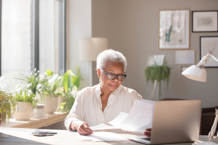 Elderly Black business woman reviewing documents on her laptop at her desk in a well-lit office, dressed in a formal blouse and glasses, the office decorated with plants and framed artwork, creating a calm, inspiring workspace for focused workの素材