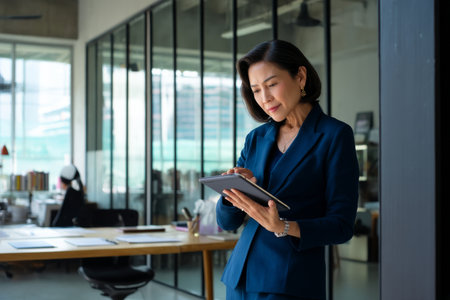 Middle-aged Asian business woman collaborating with her team in a modern office, discussing a new project over a digital tablet, dressed in a professional blue suit, the office sleek and well-organized with minimalist decor and natural light flooding in from large windowsの素材