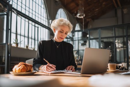 Mature Caucasian businesswoman drafting a press release, coffee and croissant beside her laptop, elegant office with high ceilings and large windowsの素材