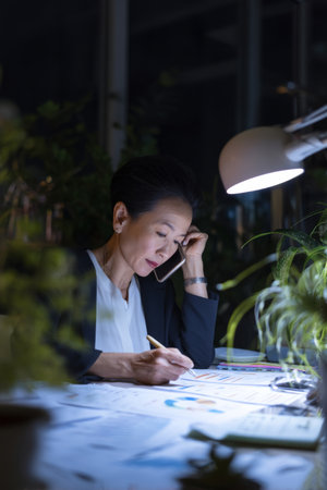 Middle-aged Asian businesswoman working late in a dimly lit office, illuminated by desk lamp, surrounded by printed analytics, phone to her ear, focused expression, plants in the background for a calming vibeの素材