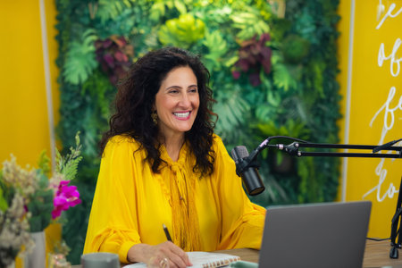 Middle-aged Hispanic business woman conducting a leadership seminar from her office, dressed in a bright yellow blouse, seated comfortably with a microphone setup, vibrant backdrop with inspirational quotes and lush greenery adds positivity to the online sessionの素材