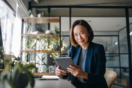 Middle-aged Asian business woman reviewing project timelines on her tablet, sitting at a modern desk in her office, dressed in a formal navy blazer, the office bright with natural light and decorated with sleek furniture and plants, creating a calm yet professional environmentの素材