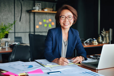 Middle-aged Asian business woman in her office, sitting at a desk covered with project documents and a laptop, engaged in a virtual meeting, wearing a navy blue suit and glasses, with a well-organized workspace in a corporate settingの素材