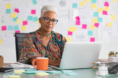 Middle-aged Black businesswoman using a laptop at her desk, wearing stylish eyeglasses, surrounded by sticky notes, coffee mug, and organizational charts on a whiteboard in the background.の素材