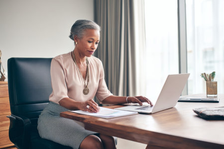 Middle-aged African-American business woman seated in her office, working on a laptop while reviewing a report, her office well-lit with minimalist decor, wearing a formal blouse and pencil skirt, exuding focus and leadershipの素材
