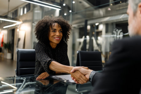 Middle-aged Black businesswoman confidently closing a deal with a handshake, seated across from a client at a glass table in a modern office with monochrome interiors and LED lighting.の素材