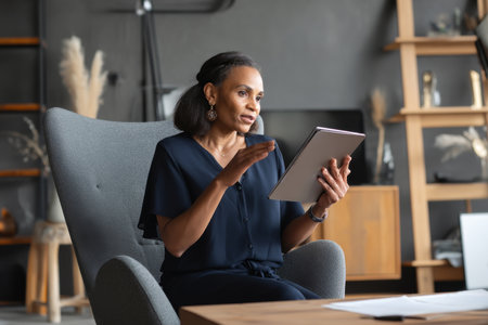 Middle-aged Black business woman conducting a virtual meeting from her office, reviewing documents on her tablet while speaking, dressed in a stylish navy blouse, her office decorated with modern furniture and minimalistic decor for a focused atmosphereの素材
