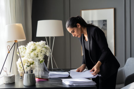 Mature Hispanic business woman working at her desk, organizing financial reports, dressed in a sleek black suit, her office neat and contemporary, the desk clutter-free with a stylish desk lamp and a vase of fresh flowersの素材