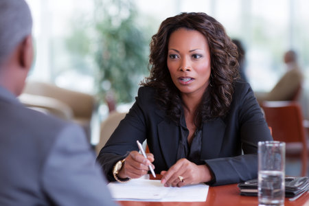 Middle-aged African-American businesswoman conducting interviews at a round table, holding a pen, reviewing resumes, and speaking with a poised and professional demeanor.の素材