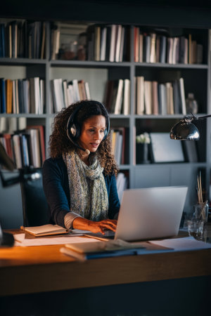 Middle-aged Black business woman video conferencing from a sleek office space, headset on, multitasking between her laptop and notes, with soft natural lighting, a tidy desk, and shelves filled with industry booksの素材