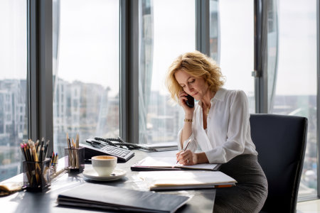 Middle-aged Caucasian business woman having a phone conversation in her office, writing notes on a notepad while sipping coffee, her workspace modern and clean, wearing a formal blouse and pencil skirt, with large windows in the backgroundの素材