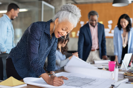 Mature Black businesswoman reviewing architectural blueprints on her desk, surrounded by team members offering feedback. The room feels dynamic, filled with creative energy and teamwork.の素材