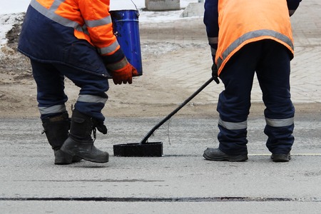 Two road workers in overalls are poured with hot tar on the edge of the road strip when repairing the roadの写真素材