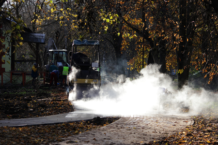 road mini building roller is working on the repair site of the bike path next to the sidewalk. white vapor glows in the sun.の写真素材