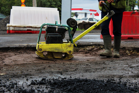 A road construction worker compacts the soil with a compact vibroplate before asphalting a...