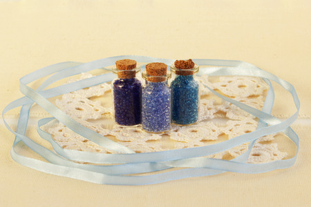 Three tiny glass bottles with a cork stopper, filled with a blue colours of beads, white knitted napkin and blue ribbon for decoration on a beige background.の写真素材