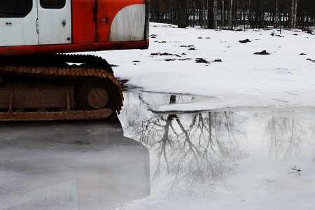 An old excavator with rusted caterpillars stands in a puddle where a tree is reflected. Spring.の写真素材