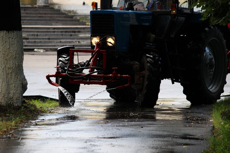 tractor with a barrel on a trailer pours water over the city sidewalks, streams of water flow down to the groundの写真素材