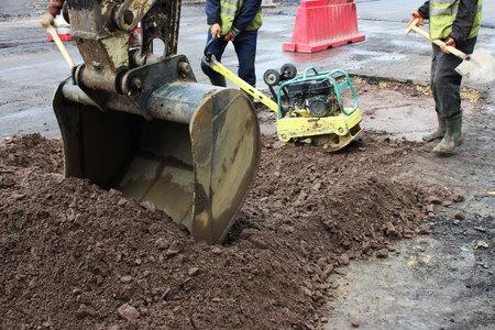 The foreman of the construction team works with a vibrating platform plate, tamping up the sand before the asphalting of the road using the patch method. reportage. foreground excavator bucket.の写真素材