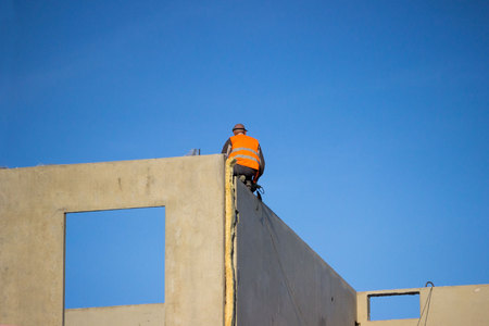 professional construction worker sits on the edge of the wall of a house under building construction, waiting for the slab, which is carried by the crane on industrial site. fifth floor. safety breach.の写真素材