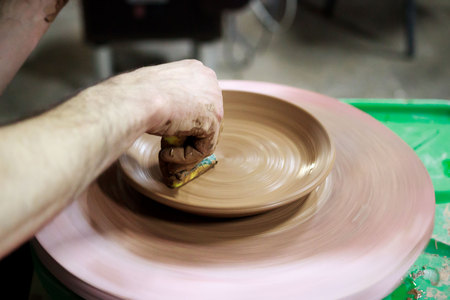 a master ceramist with 13 years of experience makes plate from red clay on a potters wheel, on a sheet of particle board for better removal and further drying. removal of excess moisture from the product using a foam sponge. reportage.の写真素材