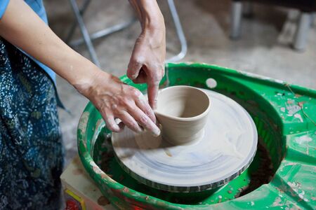 Young woman novice student in the first lesson in pottery tries to make a product from white clay on a potters wheel. reportage. small sauce pan. making a spoutの写真素材