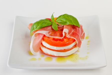 home made salad with slices of fresh mozzarella and organic tomatoes with basil and proscuitto and olive oil served on a white plateの写真素材