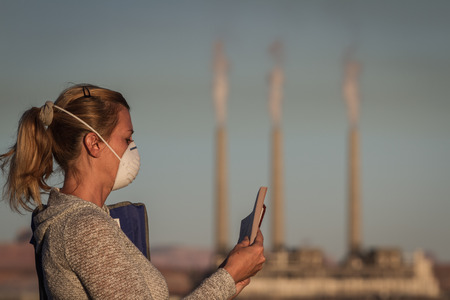 concept image of a woman sitting reading a book wearing a mask with a coal power plant and dirty smoke in the backgroundの写真素材