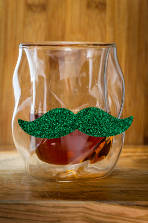 Close up of a glass of whiskey with a green mustache over a wooden background as a concept for Saint Patricks dayの写真素材