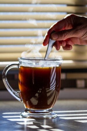 Close up of a fresh steaming cup of coffee served indoors with blinds allowing enough light to light up the steam and countertopの写真素材