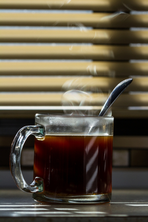 Close up of a fresh steaming cup of coffee served indoors with blinds allowing enough light to light up the steam and countertopの写真素材