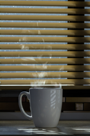 Close up of a fresh steaming cup of coffee served indoors with blinds allowing enough light to light up the steam and countertopの写真素材