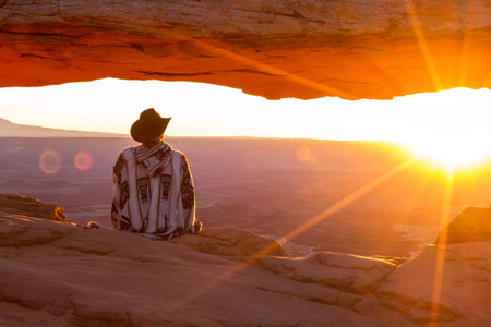 Man in western attire sitting on the edge of the cliff as the sun rises across the canyon with Mesa...