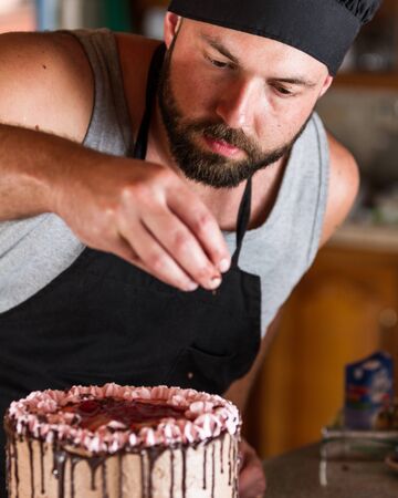 male baker decorating a chocolate birthday cake with strawberry moose frosting and dark chocolate lines on its sideの写真素材