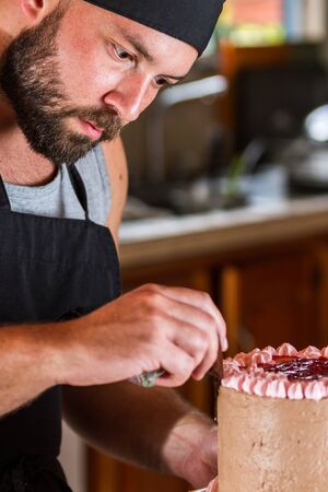 male baker decorating a chocolate birthday cake with strawberry moose frosting and dark chocolate lines on its sideの写真素材