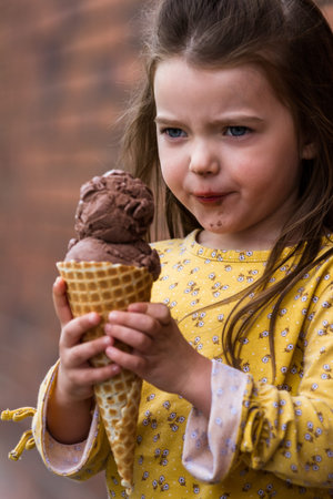 Close up of an adorable little girl eating a large chocolate ice cream cone with a brick wall in the...