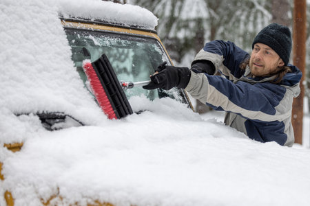 A man is clearing snow off a car window. The scene is cold and snowy, and the man is wearing a black hatの写真素材