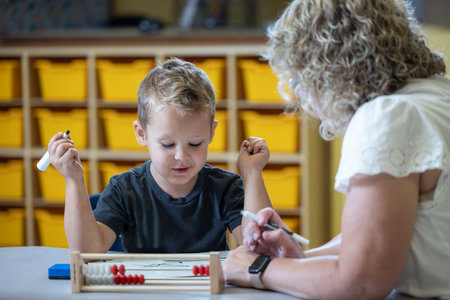 A young boy is sitting at a table with a teacher, who is helping him with a math problem. The woman is wearing glasses and has a serious expression on her face. The boy is focused on the task at handの写真素材