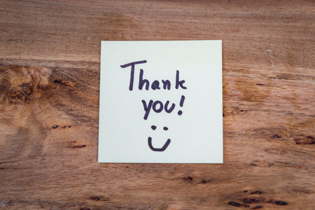 A small piece of paper with the word thank written on it. The paper is sitting on a wooden surface