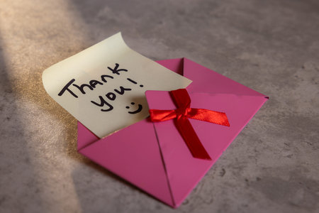A pink envelope with a red ribbon and a note that says Thank You! on it. The envelope is open and...