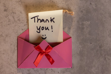 A pink envelope with a red ribbon and a note that says "Thank You!" on it. The envelope is open and the note is written on a piece of paper. Concept of gratitude and appreciationの写真素材