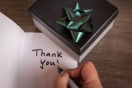 A person is writing a thank you note on a piece of paper. The note is written in black marker and next to a gift box with a ribbon.の写真素材