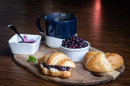A plate of croissants with huckleberry jam and a cup of coffee sit on a wooden table. The croissants are arranged on a wooden tray, and the coffee is in a blue mug. Concept of comfort and relaxationの写真素材
