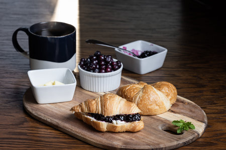 A plate of croissants with huckleberry jam and a cup of coffee sit on a wooden table. The croissants...